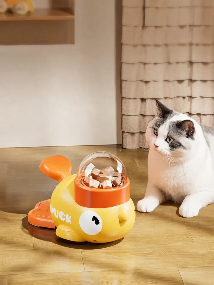 White and gray cat beside a yellow duck treat dispenser with a clear treat dome